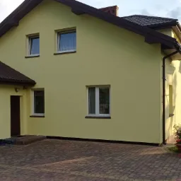 Żółty dom jednorodzinny po malowaniu elewacji, z brązowym dachem, widoczne rusztowanie z boku i kostka brukowa na podjeździe.