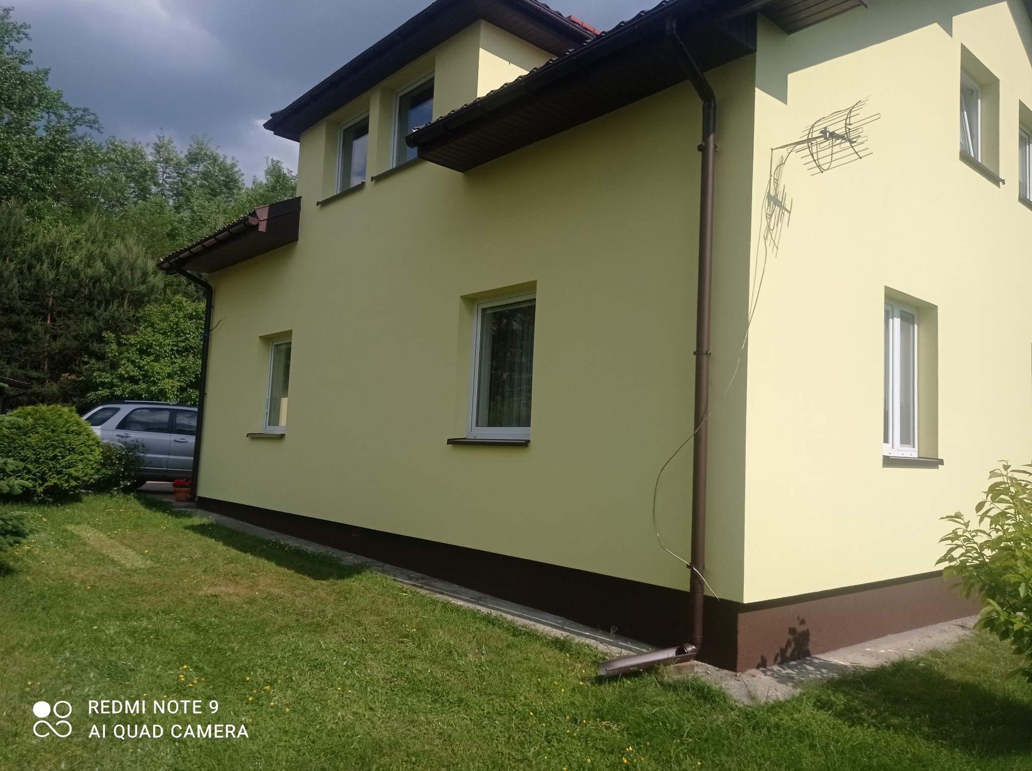 Żółta elewacja domu jednorodzinnego z brązowym cokołem, widoczna antena na ścianie, trawnik przed budynkiem i fragment samochodu w tle.