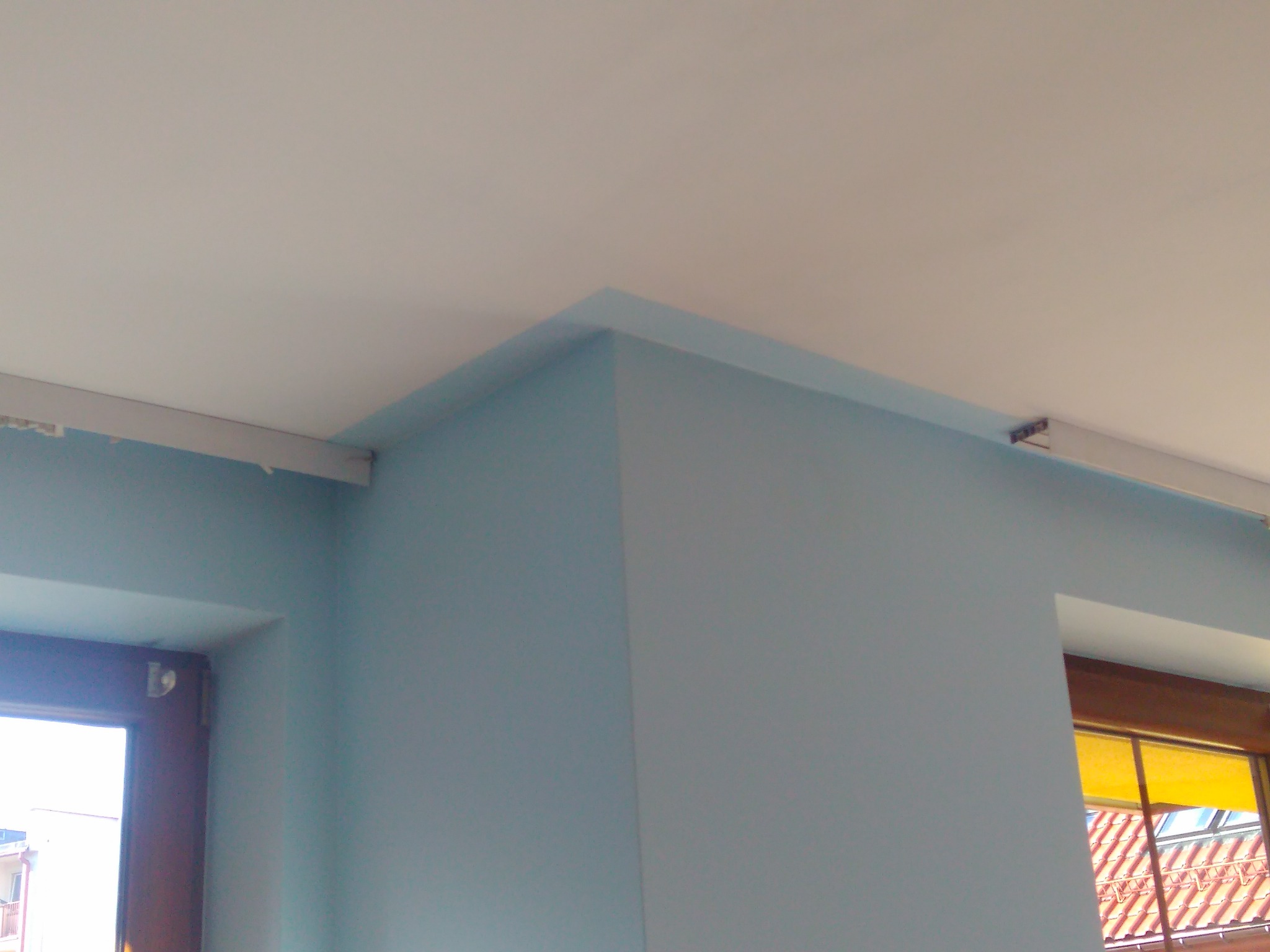 Świeżo pomalowany narożnik pokoju w odcieniach błękitu, widoczny fragment okna z roletą w kolorze żółtym i biały sufit z listwą karniszową.