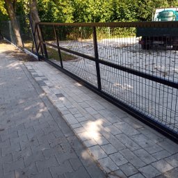 ProFence - Nowoczesne, przesuwne ogrodzenie panelowe w kolorze czarnym, zamontowane na kostce brukowej. W tle widoczny samochód dostawczy i zieleń drzew.