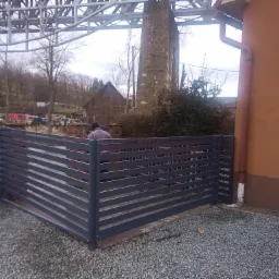 Nowoczesne, poziome, metalowe ogrodzenie panelowe w kolorze grafitowym, zamontowane na posesji z widokiem na wiadukt kolejowy i budynek mieszkalny w tle.