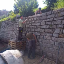 Trzech mężczyzn buduje kamienny murek oporowy, widoczna betoniarka, paleta z kamieniami i narzędzia budowlane.