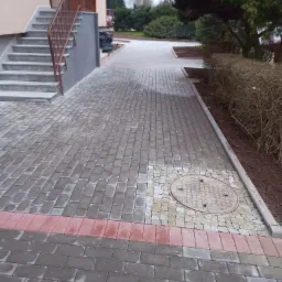 Ułożona nawierzchnia z szarej kostki brukowej w pobliżu budynku z schodami, z fragmentem czerwonej kostki i studzienką kanalizacyjną obłożoną jaśniejszymi kamieniami.