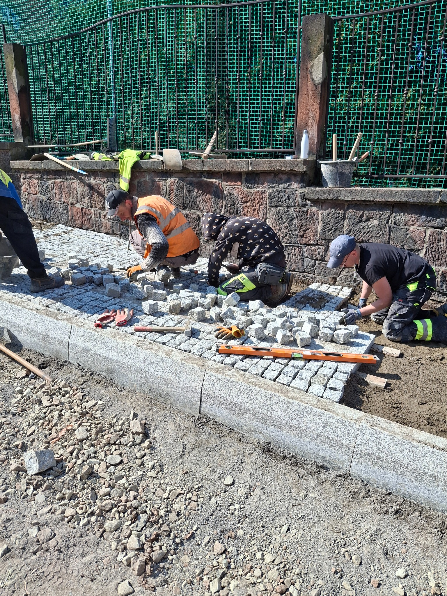 Układanie szarej kostki brukowej przez trzech pracowników w Nowej Rudzie, widoczne narzędzia i poziomica, w tle zielona siatka ogrodzeniowa.