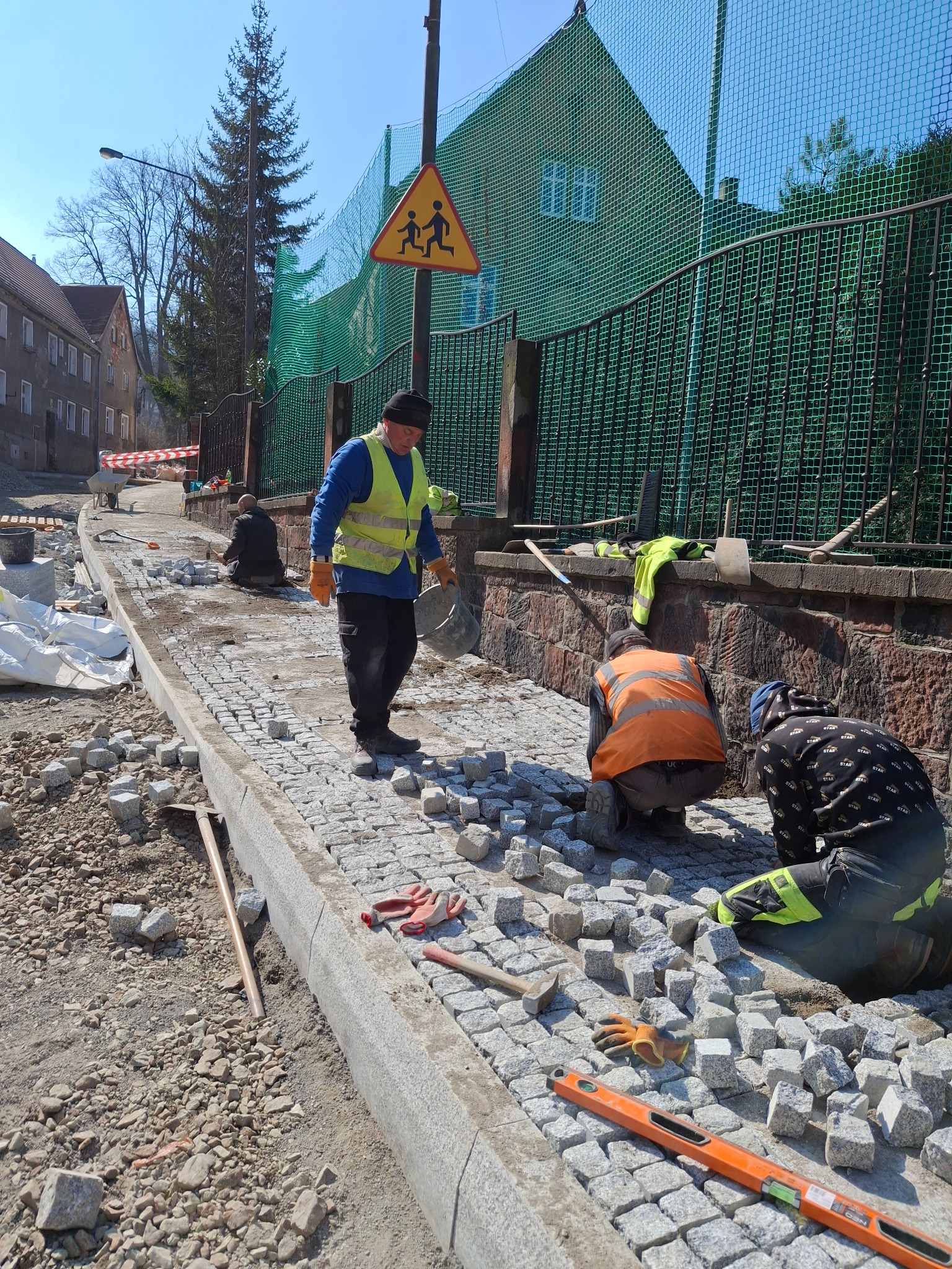 Robotnicy układają kostkę brukową na chodniku wzdłuż ogrodzenia, z widocznym znakiem ostrzegawczym dla pieszych i narzędziami pracy.