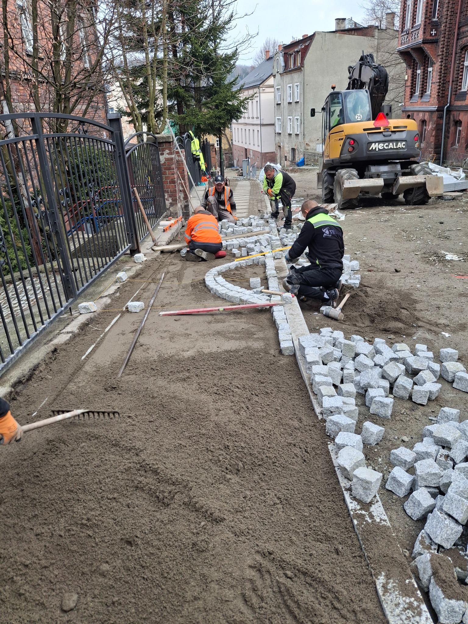 Układanie kostki brukowej z granitu na podsypce piaskowej przy użyciu poziomicy i sznurka, w tle koparka Mecalac i budynki mieszkalne.