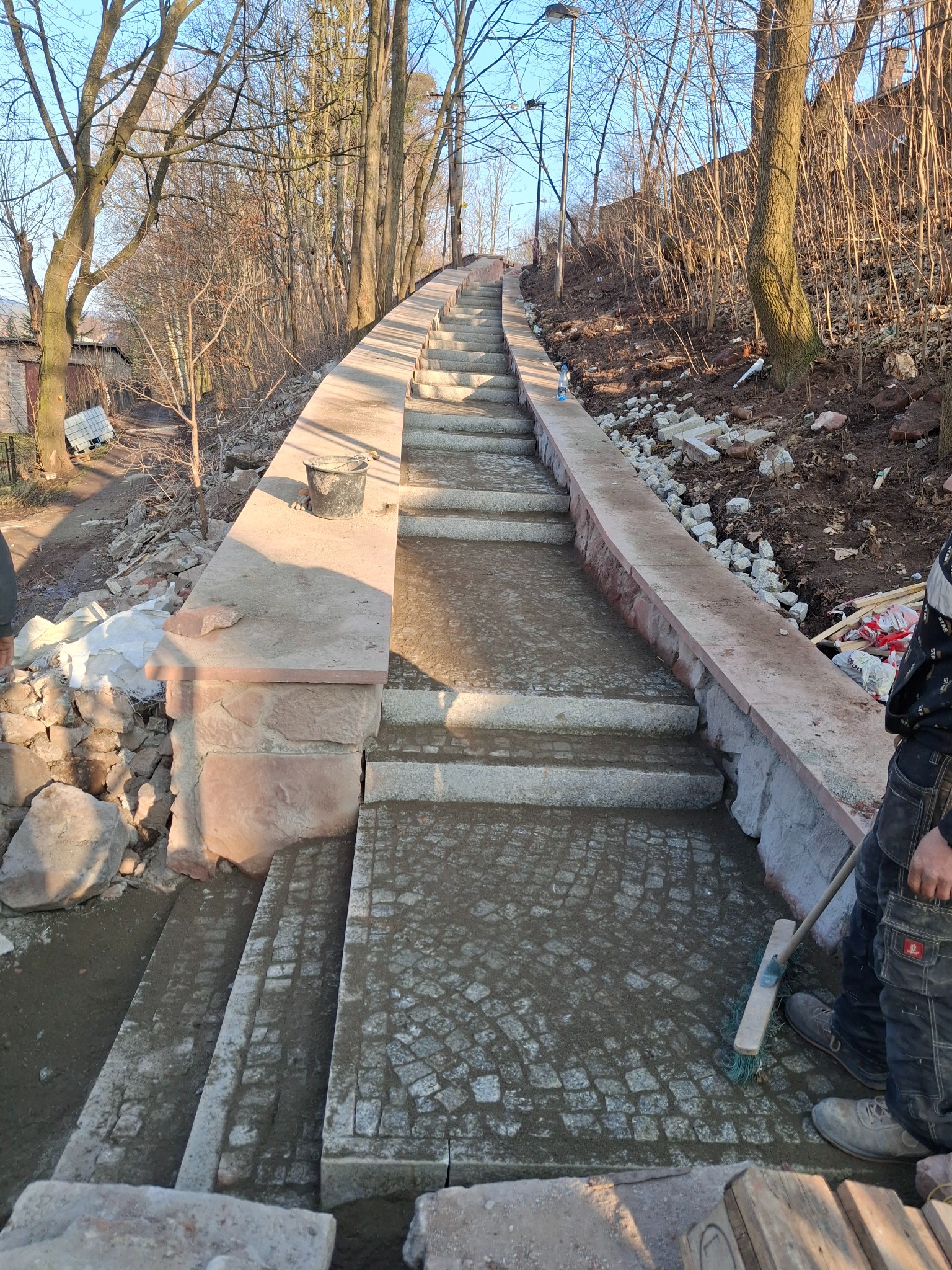 Ułożona kostka brukowa na schodach w parku, widok z góry, z boku widoczny pracownik z miotłą i wiadro z zaprawą.