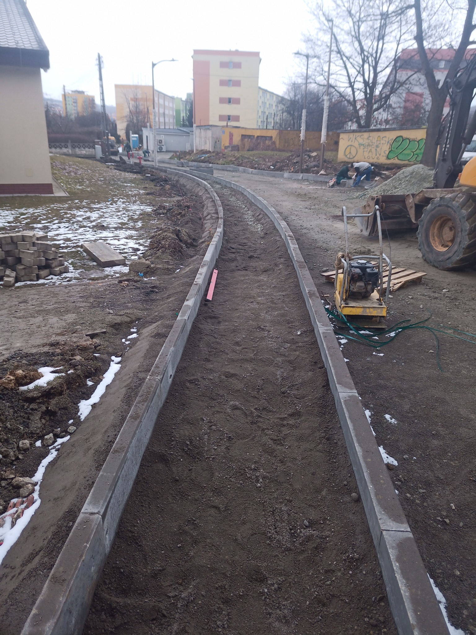 Budowa chodnika z obrzeżami betonowymi, widoczna ubijarka wibracyjna, koparka i pracownicy w tle.