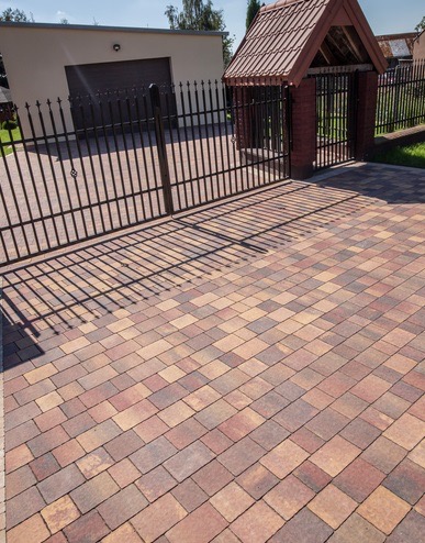 Wzór ułożonej kostki brukowej w odcieniach brązu i beżu, z metalową bramą i budynkiem w tle, widok z góry.