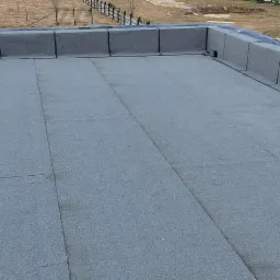 Szary dach płaski pokryty papą termozgrzewalną z wykończonymi obróbkami blacharskimi attyki, widok z góry na tle zabudowań i drzew.