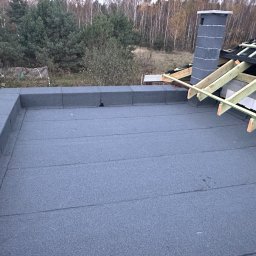 Dachpol Adam Aftyka - Płaski dach pokryty papą termozgrzewalną, z widocznym kominem i fragmentem drewnianej konstrukcji dachu skośnego w tle jesienny krajobraz.