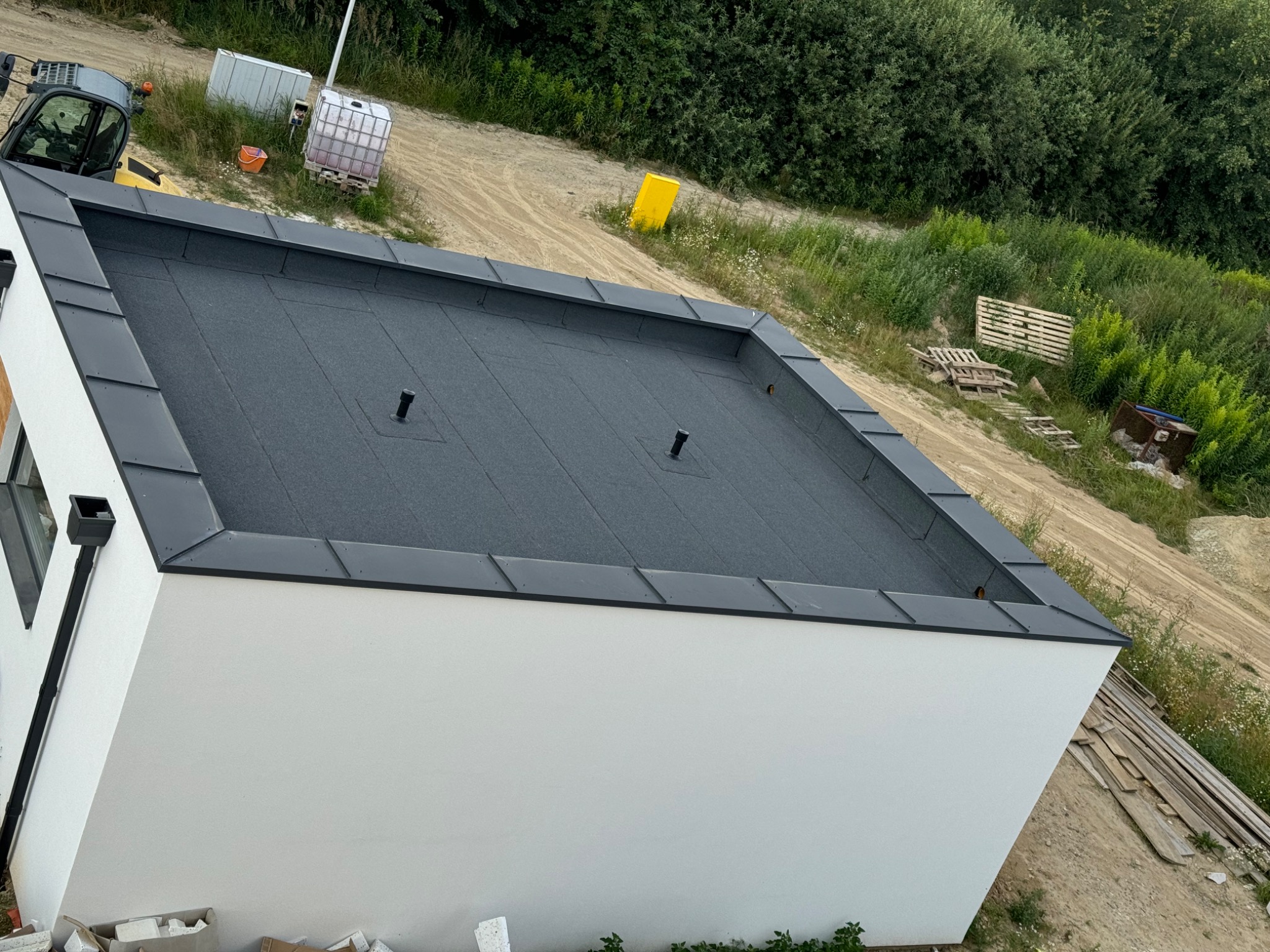 Nowoczesny, płaski dach pokryty papą termozgrzewalną z metalowymi obróbkami blacharskimi, widok z góry na biały budynek i otoczenie budowy.