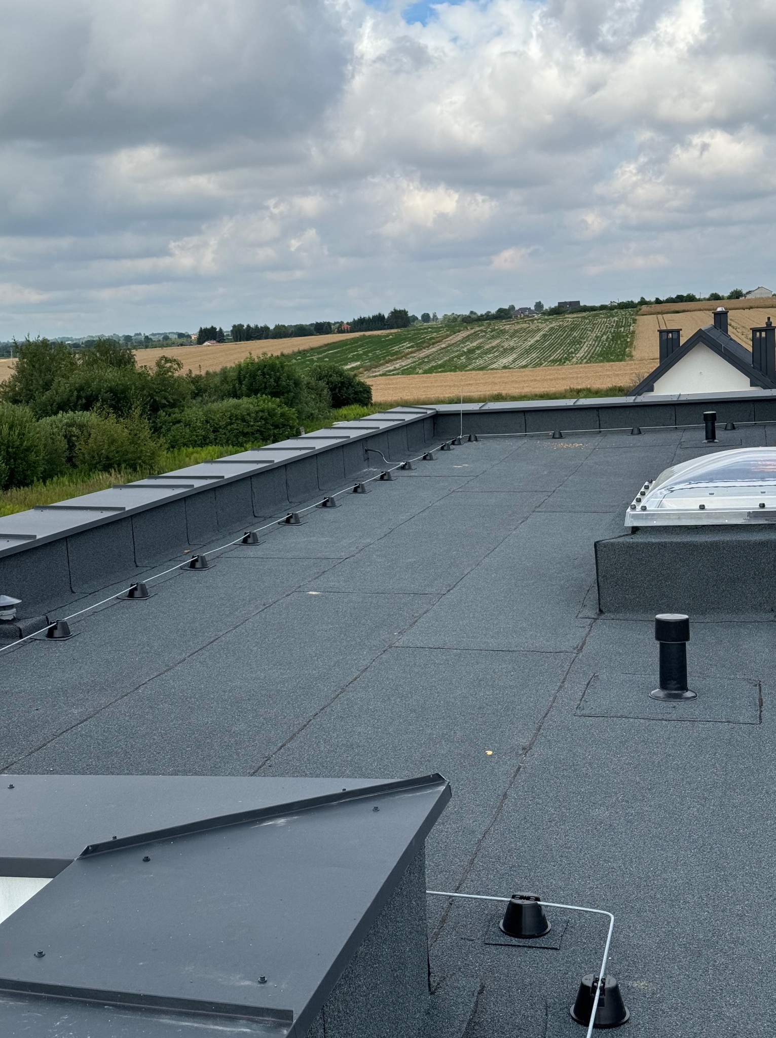 Płaski dach pokryty papą termozgrzewalną z widocznymi kominkami wentylacyjnymi, świetlikiem dachowym i systemem odgromowym na tle pól uprawnych pod pochmurnym niebem.