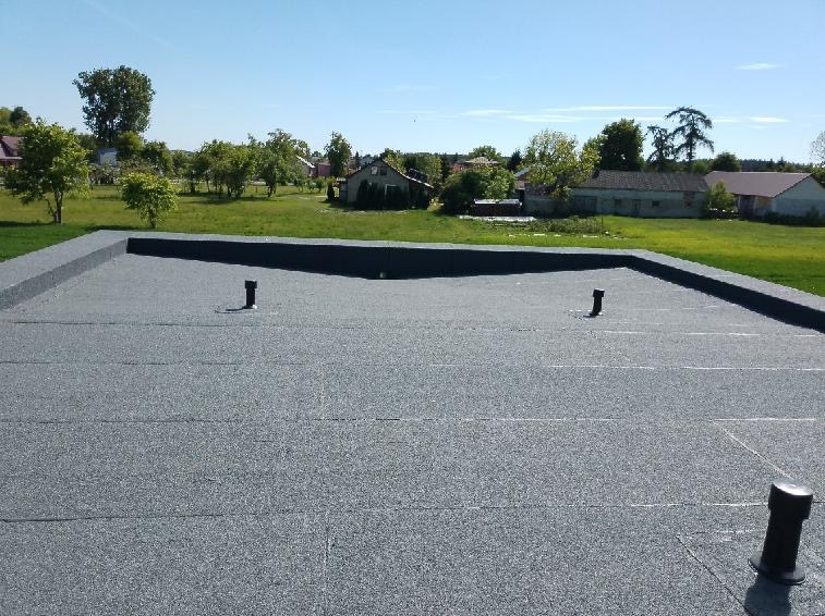 Szary, płaski dach pokryty papą termozgrzewalną z trzema kominkami wentylacyjnymi na tle zielonego krajobrazu wiejskiego.