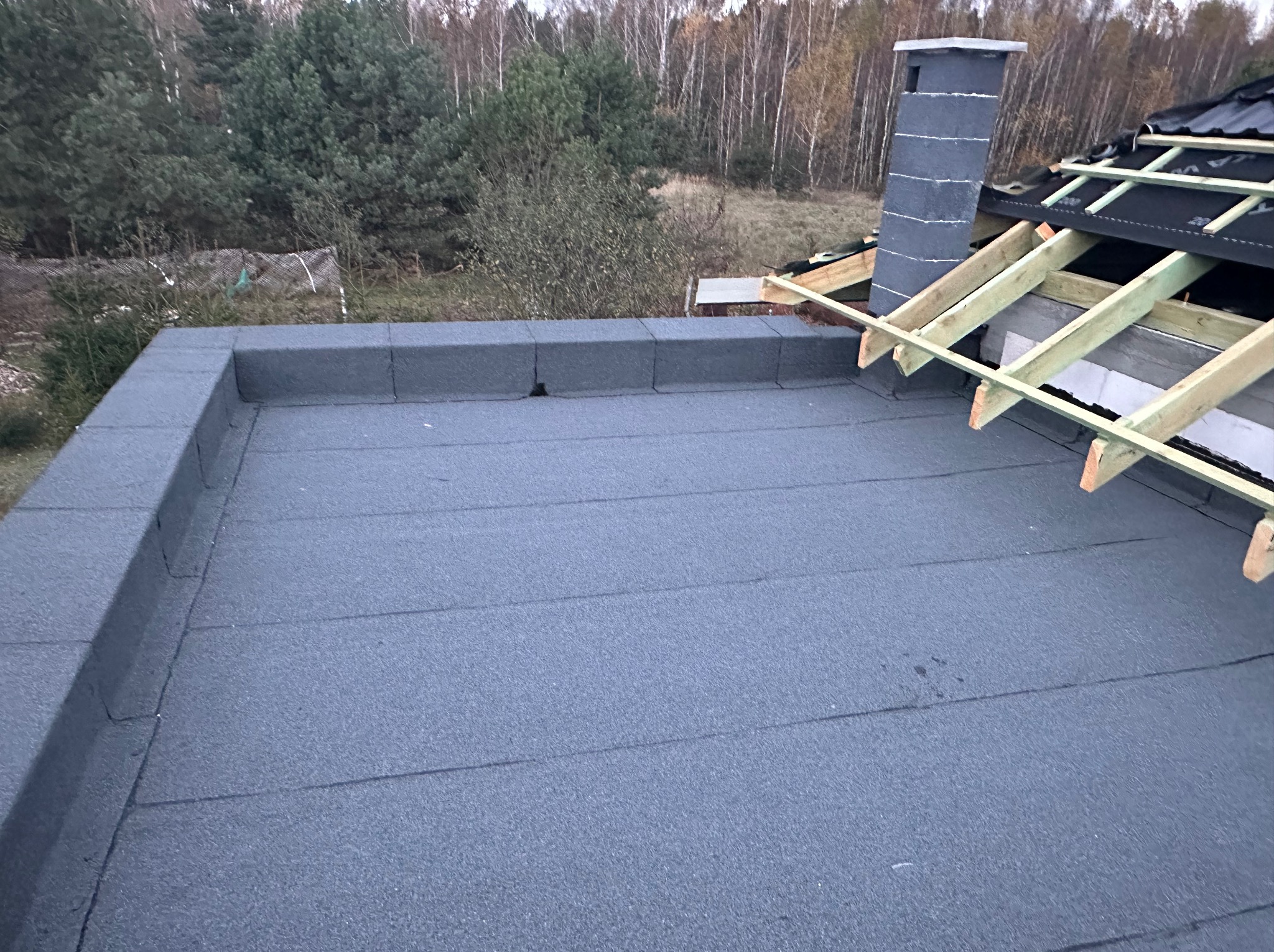 Płaski dach pokryty papą termozgrzewalną, z widocznym kominem i fragmentem drewnianej konstrukcji dachu skośnego w tle jesienny krajobraz.