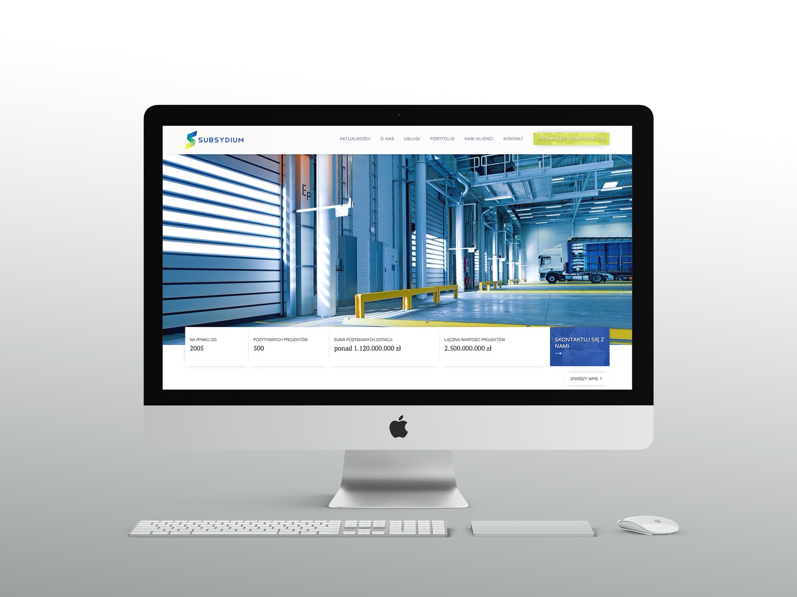 Monitor iMac wyświetlający stronę internetową firmy Subsydium z widokiem na magazyn z bramą rolowaną i ciężarówką w tle, obok klawiatura i myszka Apple.