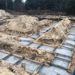 Wykop pod fundamenty z zalanym betonem i drewnianym szalunkiem, widoczne pryzmy ziemi i fragment sylwetki mężczyzny w żółtej kurtce.