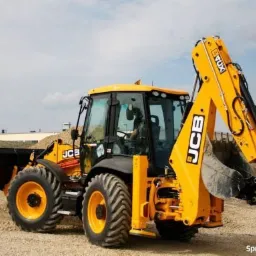 Żółta koparko-ładowarka JCB na placu budowy, z widocznym stosem ziemi w tle, opony z wyraźnym bieżnikiem, ramię koparki uniesione.