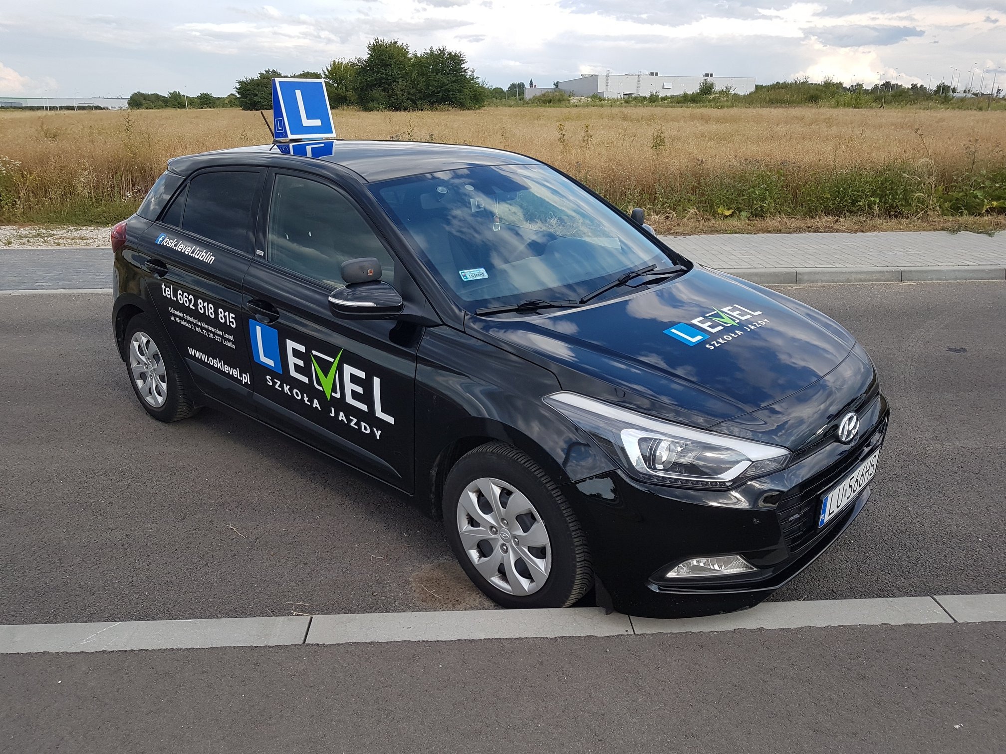 Czarny samochód z oznaczeniem 'L' na dachu, zaparkowany na poboczu drogi, z widocznym logo i danymi kontaktowymi ośrodka szkolenia kierowców Level.