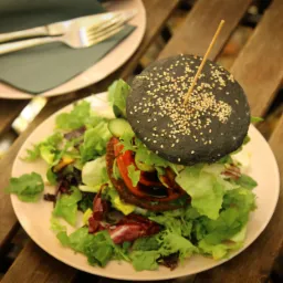 Czarny burger wegański z sezamem na talerzu z sałatą, pomidorem i ogórkiem, obok talerz ze sztućcami na drewnianym stole.