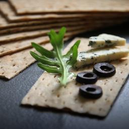 workshop44 - Krakers z serem pleśniowym, oliwkami i rukolą na ciemnym tle, ułożony obok stosu innych krakersów.