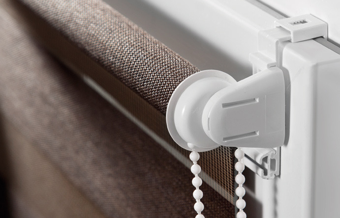 Fragment brązowej rolety okiennej z białym mechanizmem łańcuszkowym, widoczne detale tkaniny i mocowania.