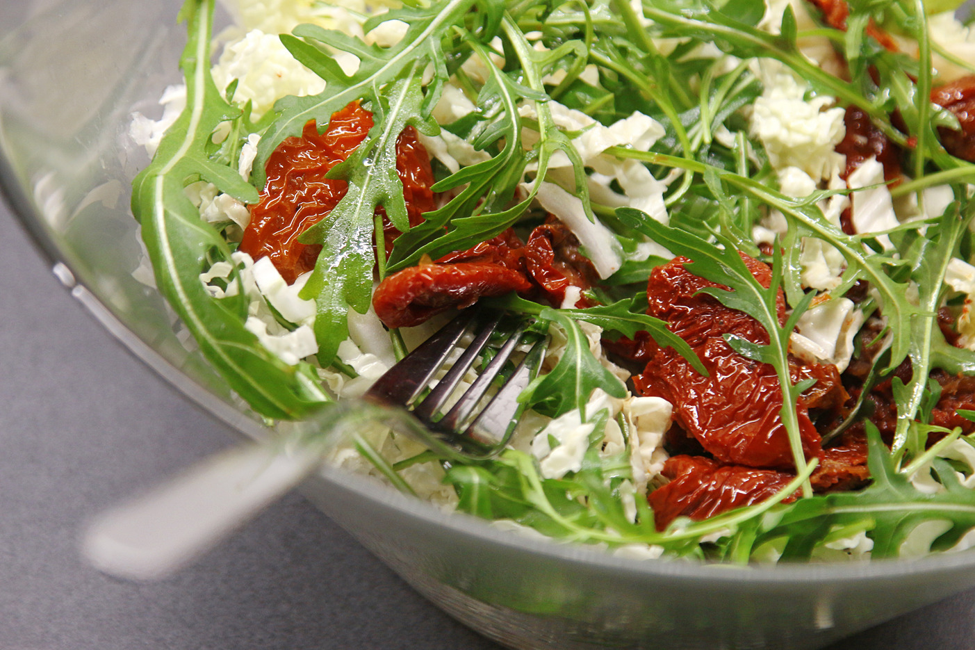 Sałatka z rukoli, suszonych pomidorów i kapusty pekińskiej w szklanej misce, z widocznym srebrnym widelcem.