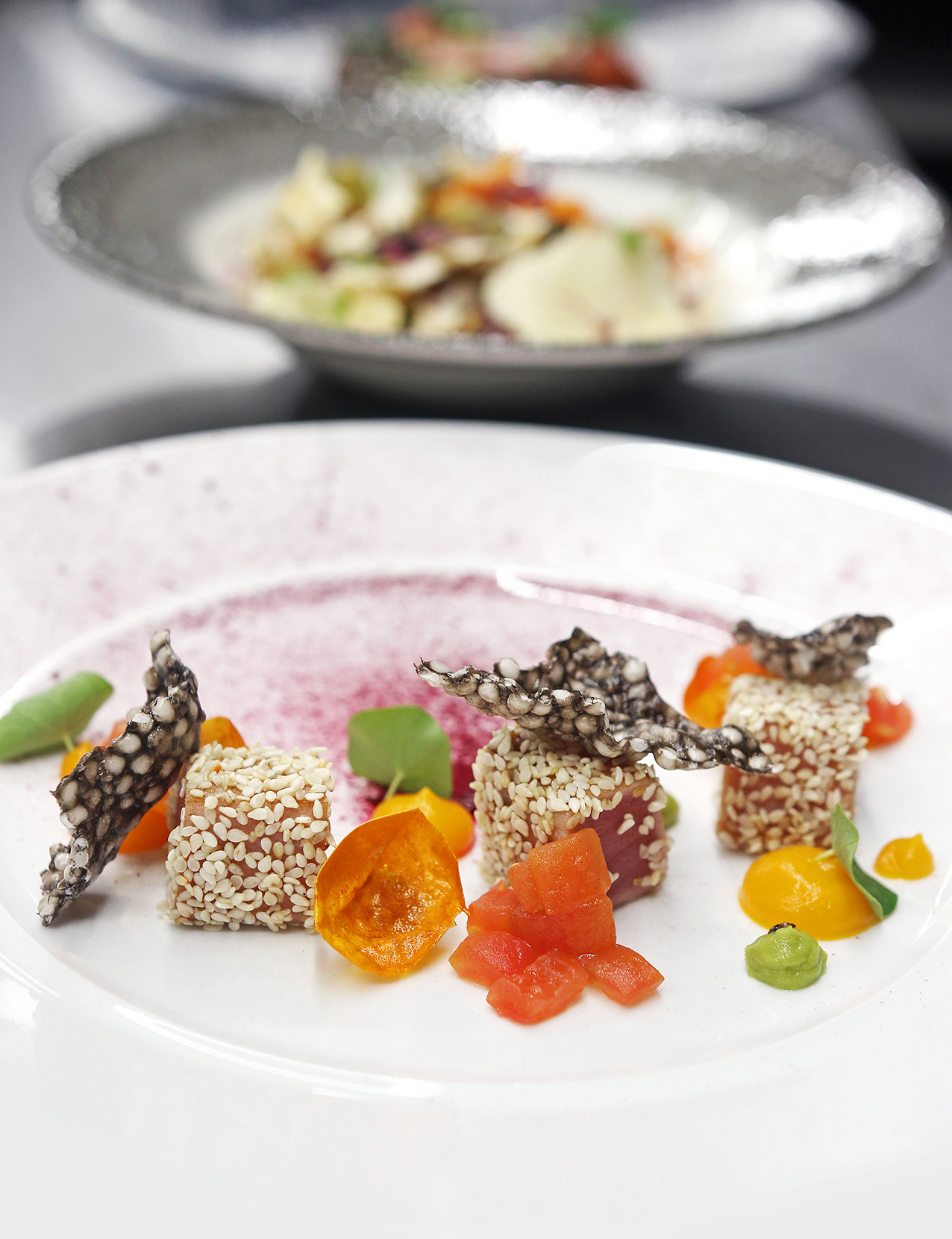 Elegancko podane danie z tuńczyka obtoczonego w sezamie, z dodatkami warzyw i jadalną dekoracją na białym talerzu z fioletową obwódką, w tle talerz z sałatką.