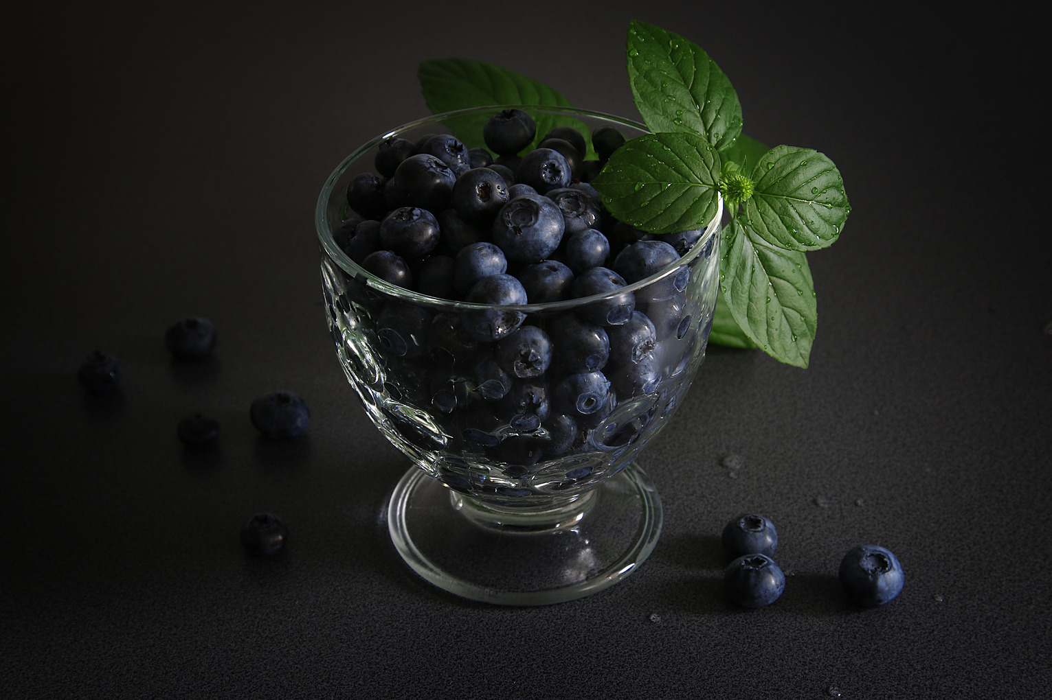 Szklany pucharek wypełniony dojrzałymi borówkami, udekorowany świeżymi listkami mięty na ciemnym tle.