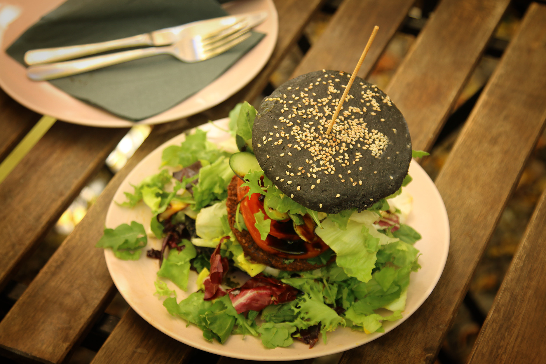 Czarny burger wegański z sezamem na talerzu z sałatą, pomidorem i ogórkiem, obok talerz ze sztućcami na drewnianym stole.