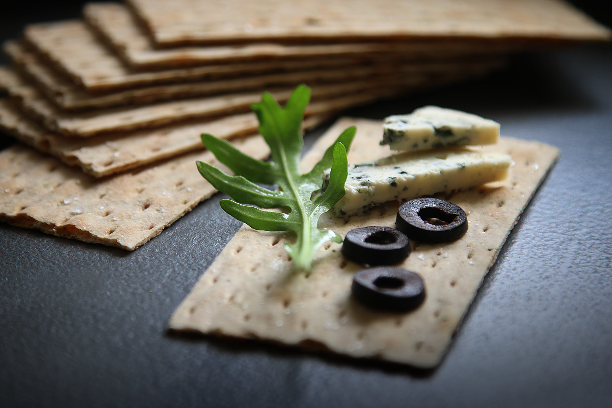 Krakers z serem pleśniowym, oliwkami i rukolą na ciemnym tle, ułożony obok stosu innych krakersów.