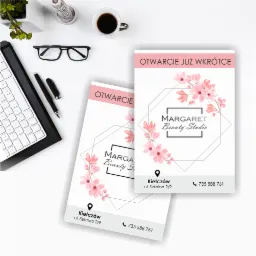 Dwa identyczne projekty graficzne, prawdopodobnie ulotki reklamowe salonu kosmetycznego 'Margaret Beauty Studio', z motywem kwiatowym i hasłem 'Otwarcie już wkrótce', leżące na białym biurku obok...