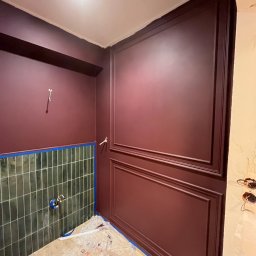 RS BUD - Świeżo pomalowane ściany w odcieniach burgundu z dekoracyjnymi panelami, dolna część ściany wyłożona zielonymi płytkami, podłoga zabezpieczona folią malarską.