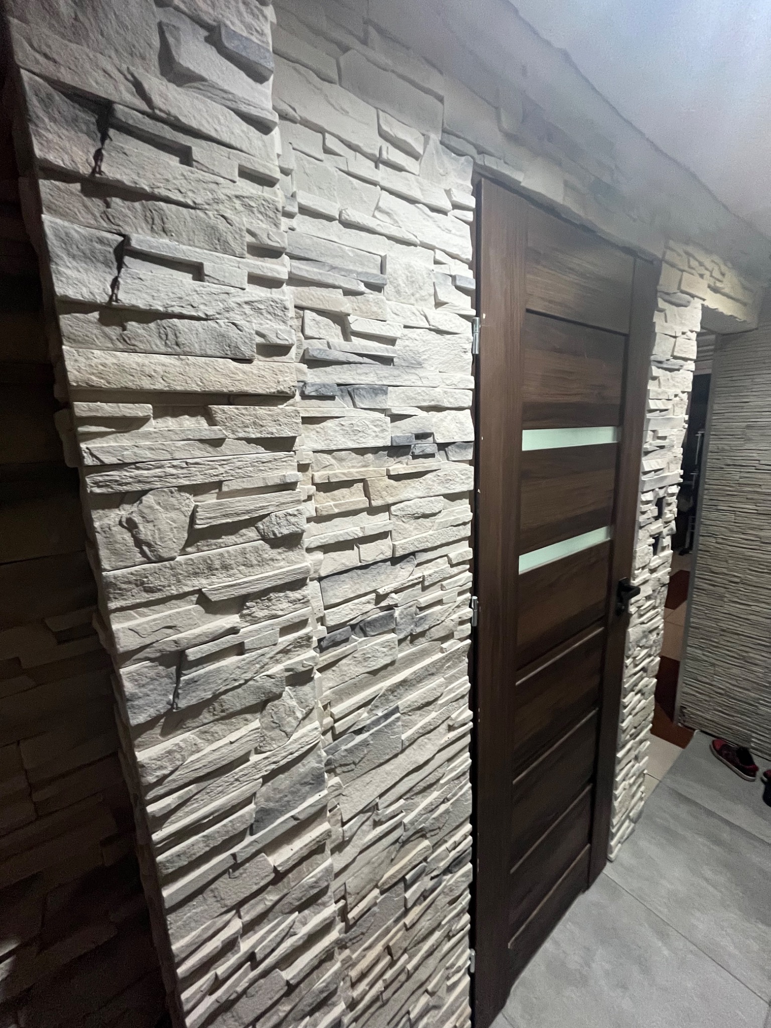 Dekoracyjna ściana z kamienia elewacyjnego w odcieniach szarości, przylegająca do drewnianych drzwi z trzema poziomymi przeszkleniami, widok na fragment podłogi i dalszą część ściany z podobnym...