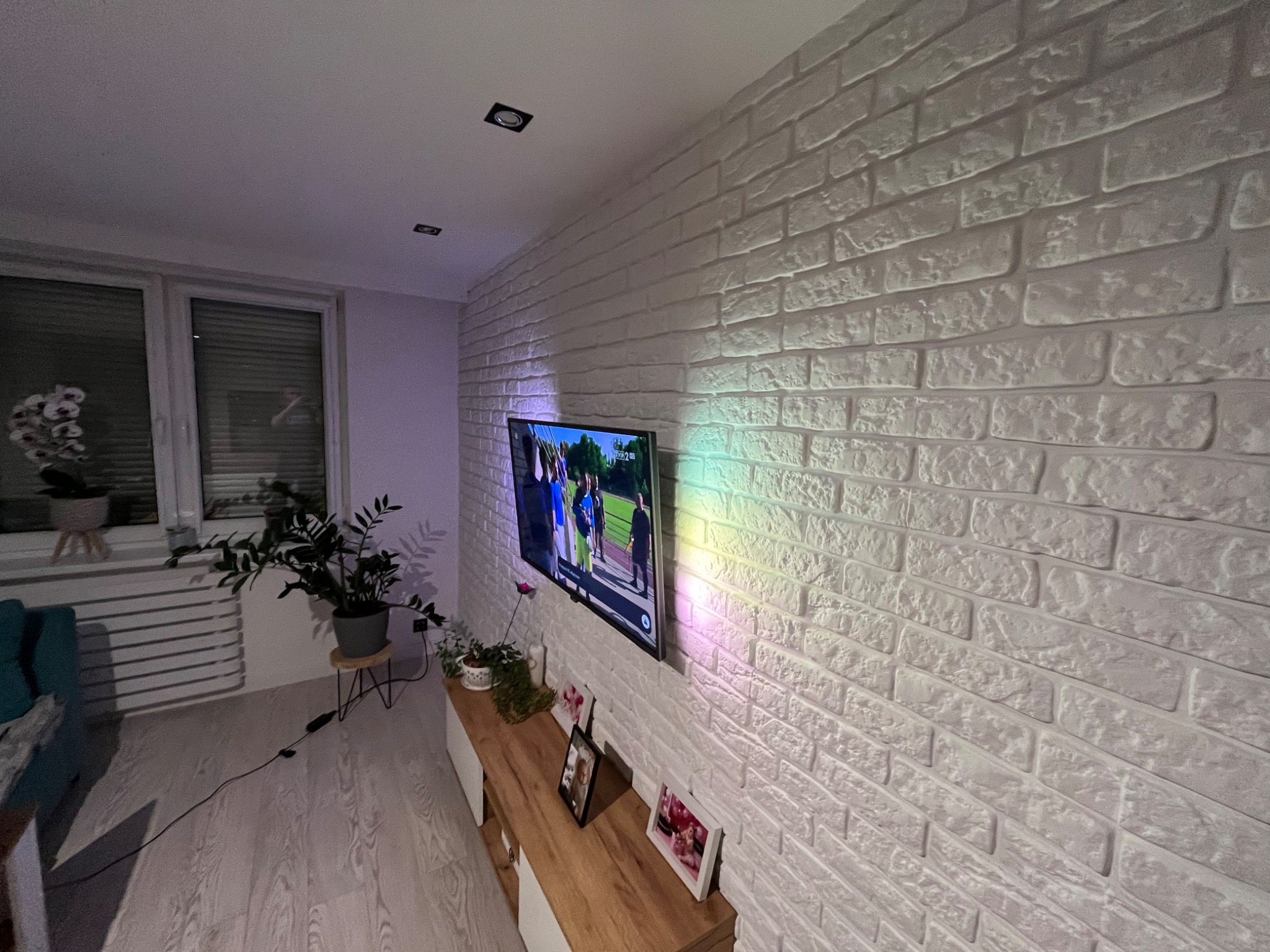 Salon z białą ścianą z cegły dekoracyjnej, telewizorem na ścianie, drewnianą komodą, roślinami w doniczkach i oświetleniem punktowym.