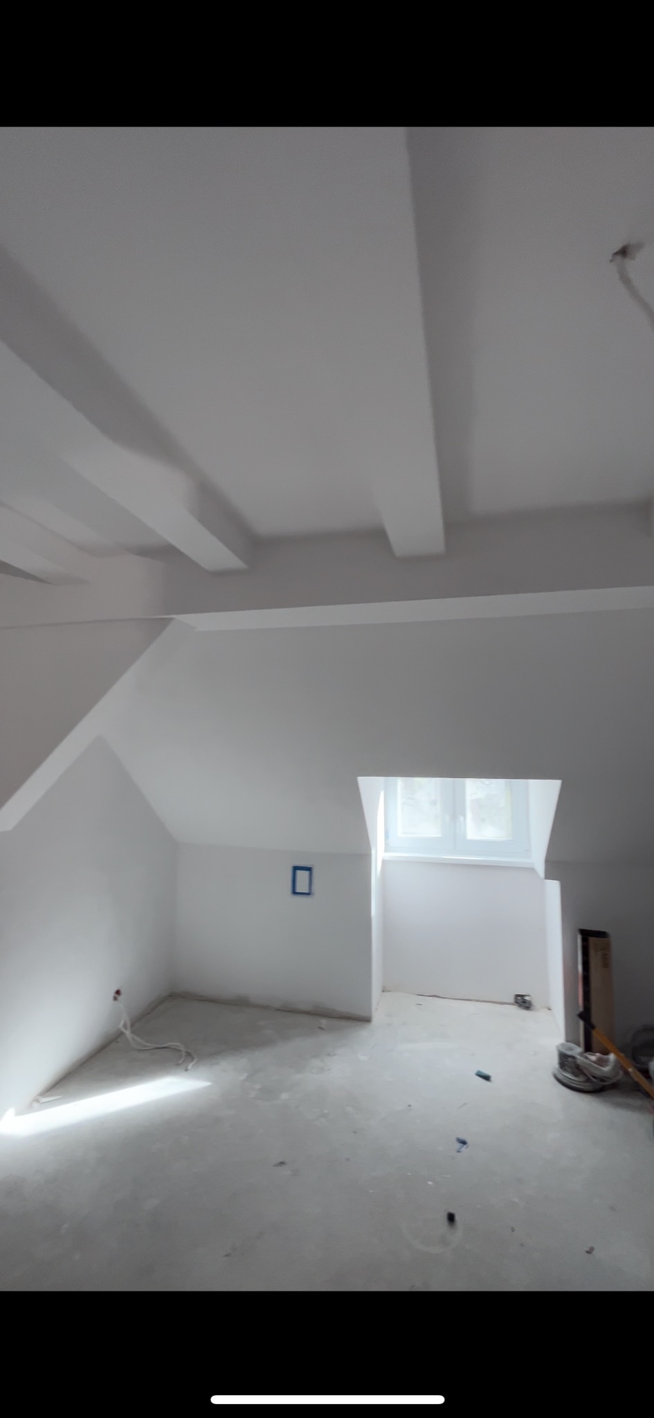 Poddasze w trakcie prac wykończeniowych: świeżo pomalowane na biało ściany i sufit z widocznymi belkami, okno dachowe w głębi, wystające kable elektryczne, podłoga przygotowana do dalszych prac.