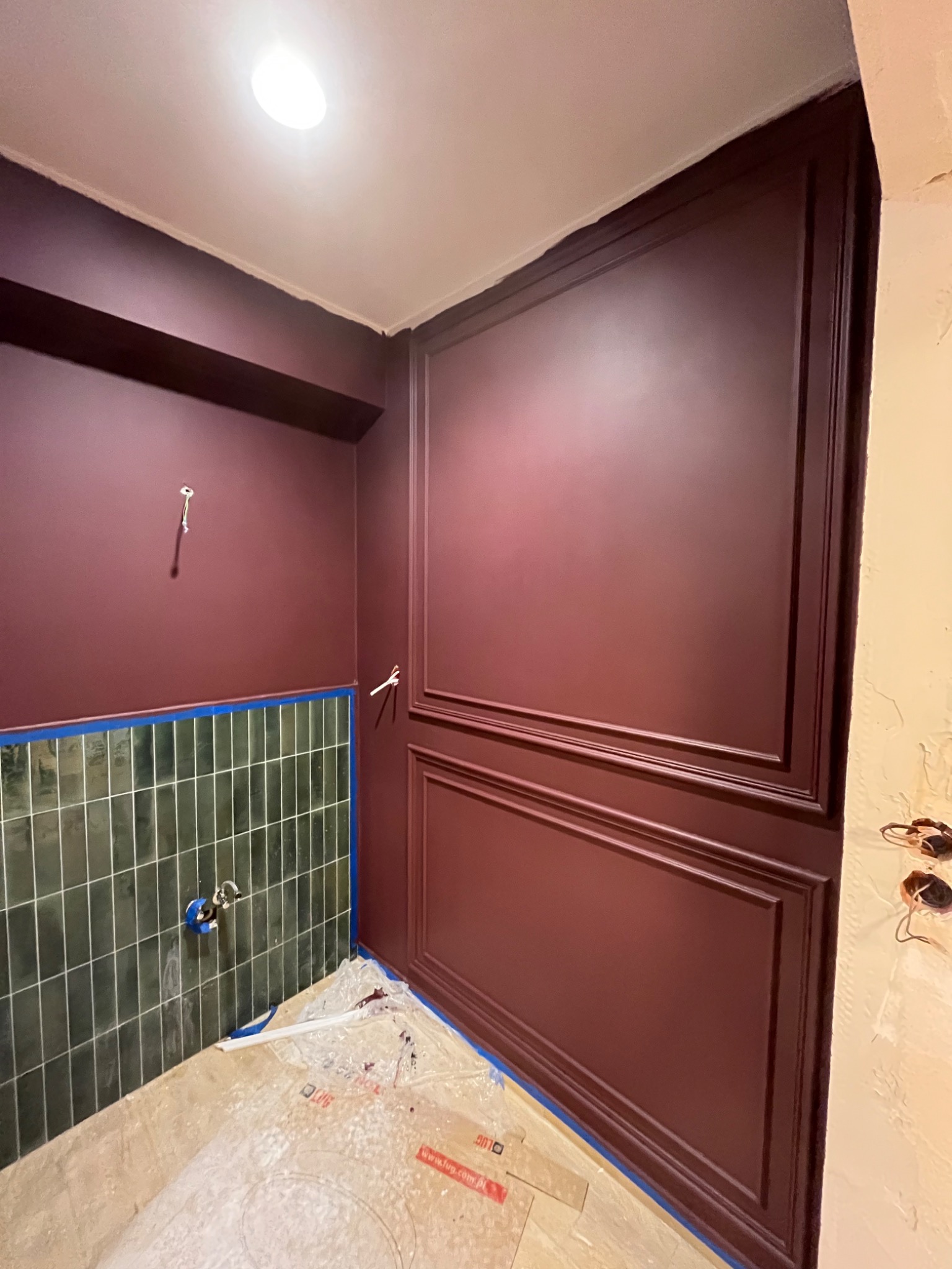 Świeżo pomalowane ściany w odcieniach burgundu z dekoracyjnymi panelami, dolna część ściany wyłożona zielonymi płytkami, podłoga zabezpieczona folią malarską.