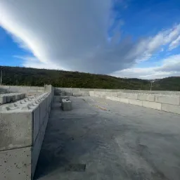 Konstrukcja z betonowych bloków modułowych tworząca mury oporowe na tle zielonego wzgórza i błękitnego nieba z chmurami.