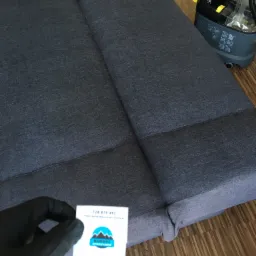 Szara, rozłożona kanapa z widocznym szwem, obok odkurzacz piorący z żółtymi elementami. W lewym dolnym rogu dłoń w czarnej rękawiczce trzyma wizytówkę firmy sprzątającej.