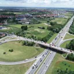 Panorama miasta z lotu ptaka, widok na autostradę z wiaduktem, otoczoną zielenią i zabudową miejską w słoneczny dzień.