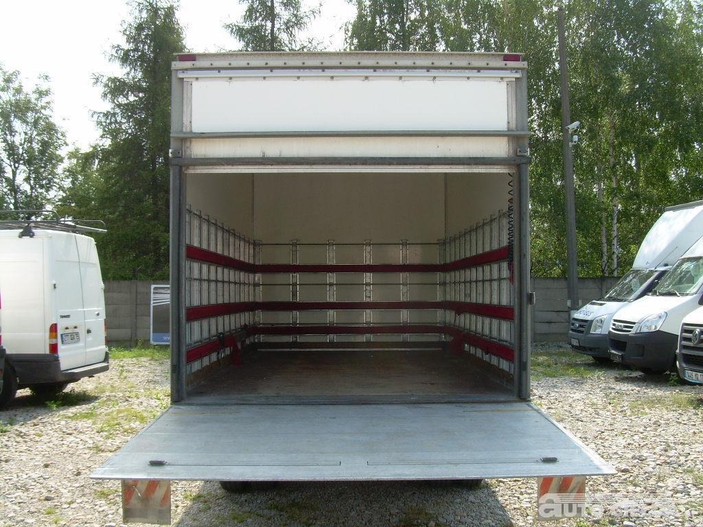 Otwarta pusta przestrzeń ładunkowa samochodu dostawczego z zabezpieczeniami ładunku, widok z tyłu na tle zieleni i innych pojazdów.