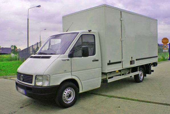 Biały samochód dostawczy z zamkniętą zabudową kontenerową, zaparkowany na betonowym placu, widoczny znak drogowy w tle.