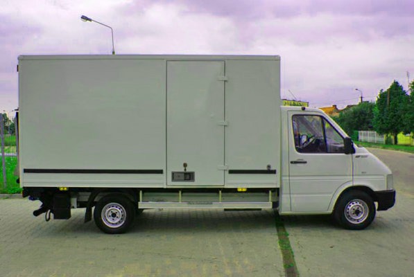 Biały samochód dostawczy z zabudową kontenerową, zaparkowany na betonowym placu, widok z boku.