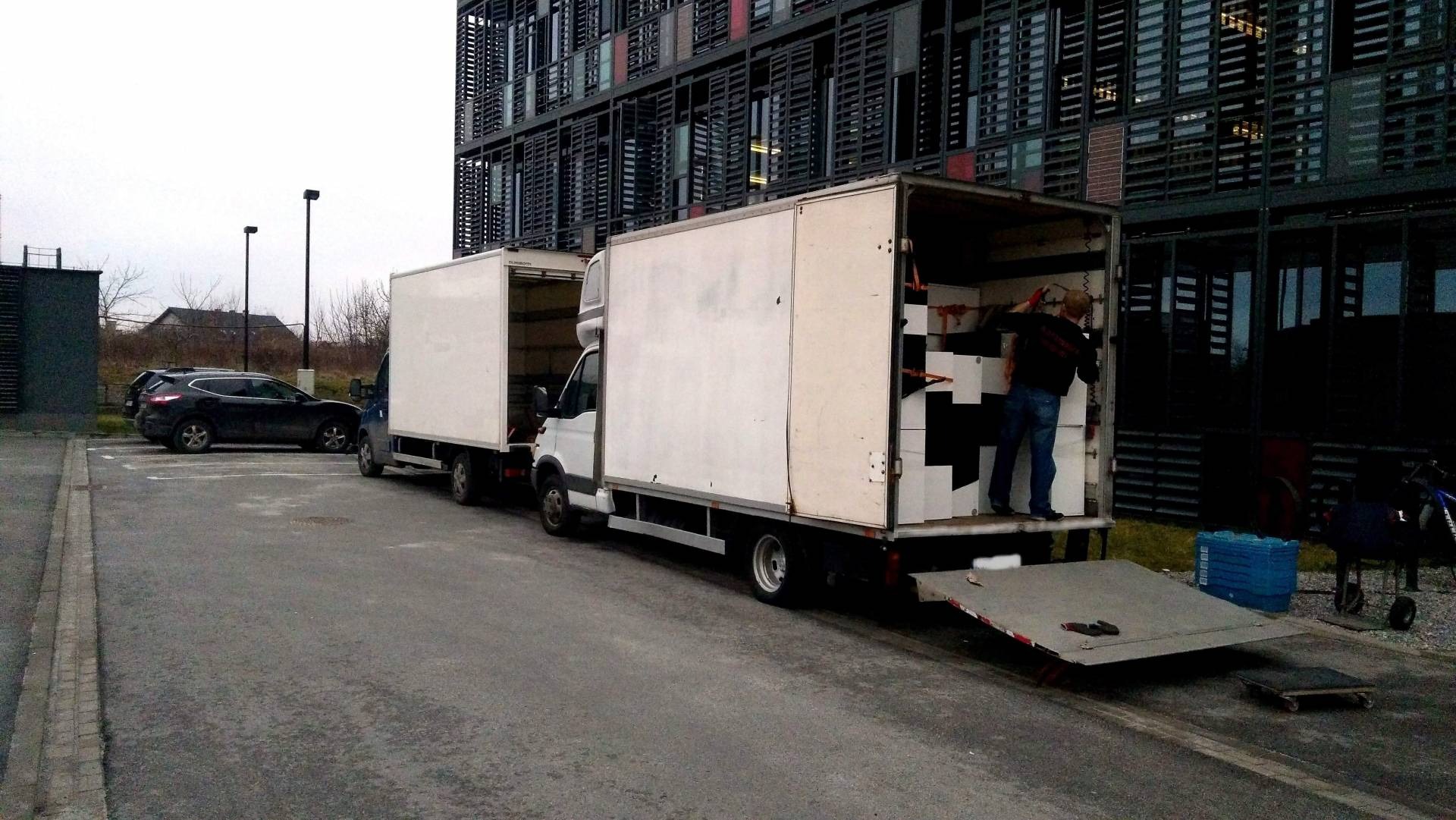 Pracownik wewnątrz naczepy białego samochodu dostawczego, układa kartony z czarnymi znakami na tle nowoczesnego biurowca z ciemną elewacją.