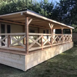 Zadaszony drewniany taras z balustradą w kształcie krzyży przy domku mobilnym, widok z zewnątrz, na trawniku.