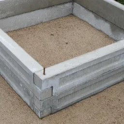 Podwyższona grządka warzywna wykonana z prefabrykowanych betonowych elementów, wypełniona piaskiem, z wystającym metalowym prętem łączącym.