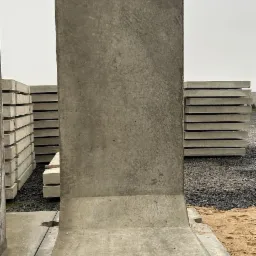 Betonowy mur oporowy typu L z dwoma stalowymi uchwytami do transportu, ułożony na stosie podobnych elementów na placu budowy.