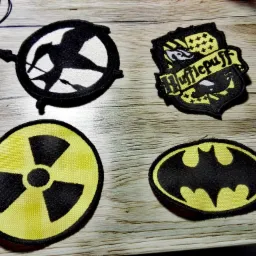 Cztery naszywki: logo Igrzysk Śmierci, herb Hufflepuff, symbol radioaktywności i logo Batmana, leżą na drewnianym blacie.