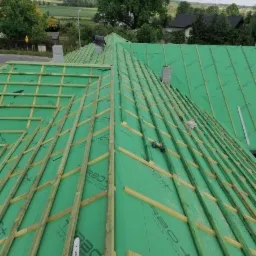 Konstrukcja dachu pokryta zieloną membraną dachową, widoczne drewniane łaty, piła spalinowa Stihl oraz wkrętarka, w tle krajobraz.