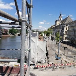 D.K.W. Inwest Ihor Kuchirka Spółka Komandytowa - Budowa lub renowacja budynku w mieście, widok z rusztowania na plac budowy, rzekę i zabudowę miejską w tle.